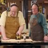Two older men using a table saw in a workshop wearing safety gear and aprons.